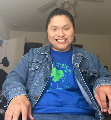 Alexa is a Latina woman with black hair who is sitting in a wheelchair wearing a blue jean jacket. She has a blue t-shirt with green lettering and a green heart in the middle that reads “cerebral palsy awareness month.”