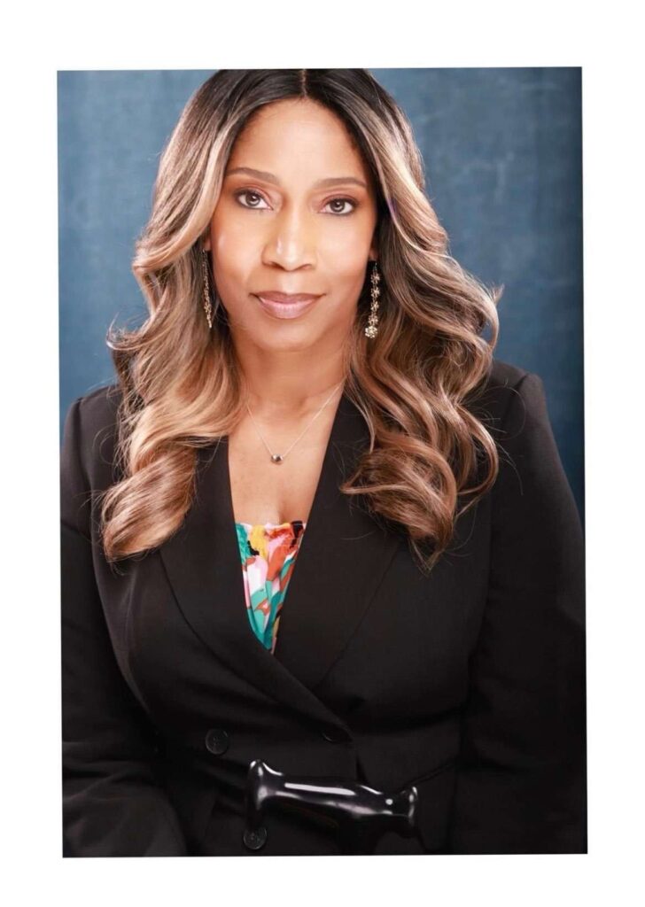 A Headshot of a Black woman with brown hair and blonde highlights is wearing a black blazer with a colorful turquoise, orange, and peach blouse.
