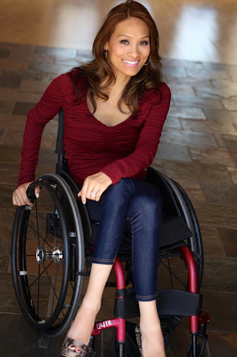 An Asian-American woman sits cross-legged in a manual wheelchair. She is smiling and wearing a long-sleeve red shirt with jeans. She has long black hair
