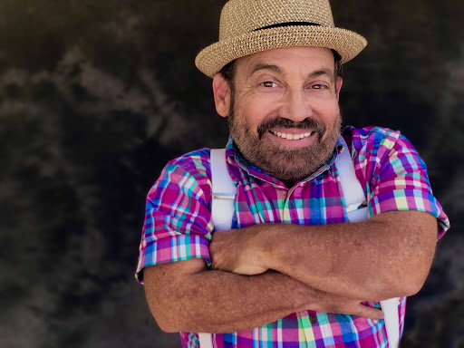 A Caucasian man with a brown goatee, wears a colorful button-down shirt, and a hat with a badass expression glares into the camera