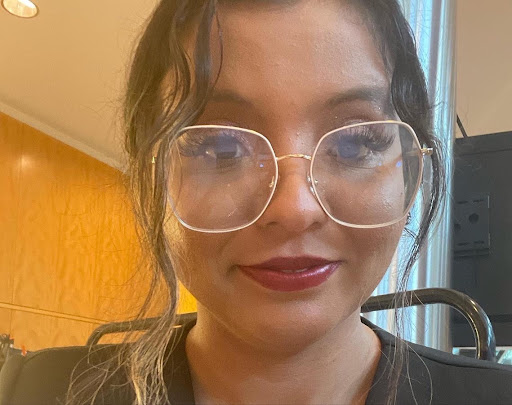 Jessica Riestra Latine youth organizer with brown skin, brown hair, brown eyes wearing glasses, and red lipstick. Sitting down smiling while wearing a black dress