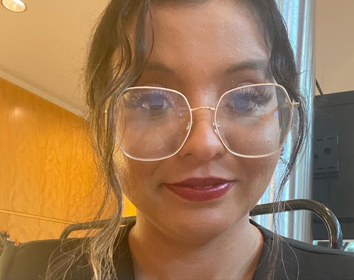 Latine youth organizer with brown skin, brown hair, brown eyes wearing glasses, and red lipstick. Sitting down smiling while wearing a black dress