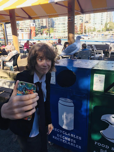 Pictured is Liz, a Caucasian, non-binary individual with light brown hair and blue eyes wearing a dark suit. Liz is holding her phone to take a selfie with an unamused seagull.