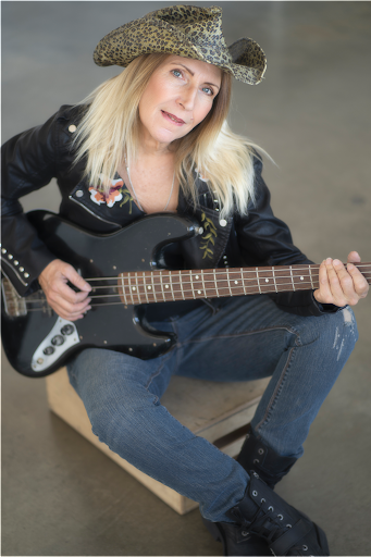Blonde caucasian woman wearing jeans, a black, flowered leather jacket, and a leopard print Western hat. She is holding a black electric bass guitar