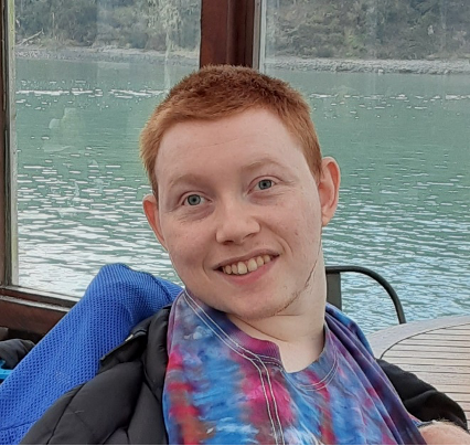 Male with red hair and tie dye shirt smiles with the background of a lake and trees behind him.