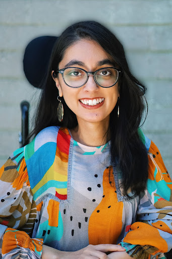 A South Asian woman with long black hair, and black glasses, wearing a multi-colored blouse against a gray wall smiles at the camera