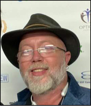 Man with beard smiling wearing blue coat, glasses and a dark brown fedora hat.