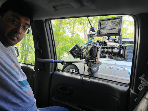 Mrigesh is sitting behind a camera. He is wearing a gray shirt with a picture of a mountain, lake, and clouds.