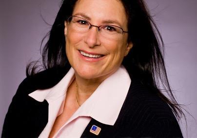 A woman with long brown hair in a pink blouse and dark jacket smiles.